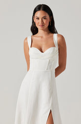 A woman is wearing a sleeveless white dress with wide shoulder straps, a sweetheart neckline with ruched detailing on the bust, and a fitted waist with vertical seam accents. The dress has a front slit on the side.
